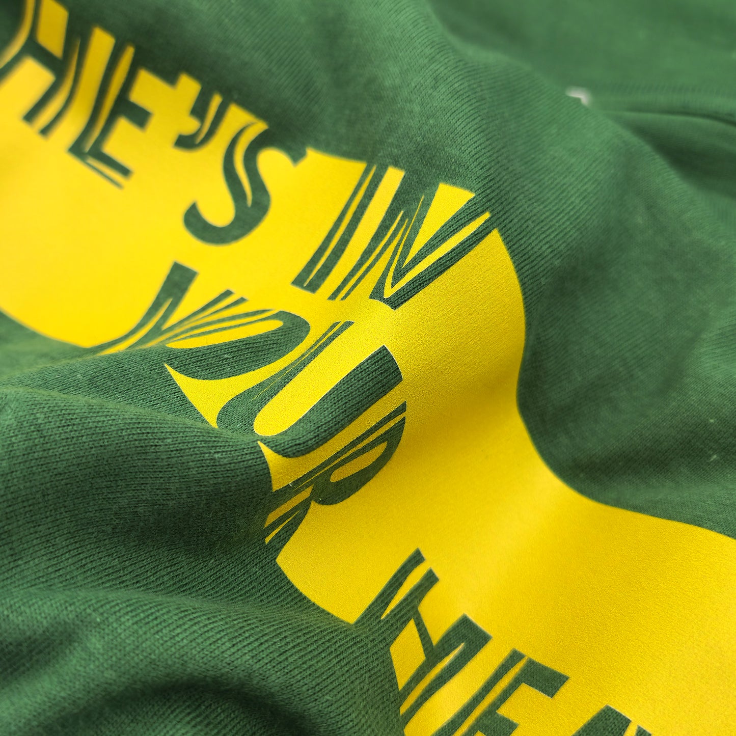 Close-up of a green t shirt with the text "He's in Your head" in yellow showing the print quality

